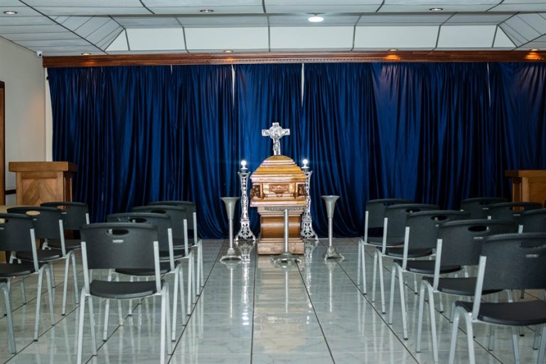 Sala de velación con ataúd, cruz cristiana, cortinas azules y sillas para familiares
